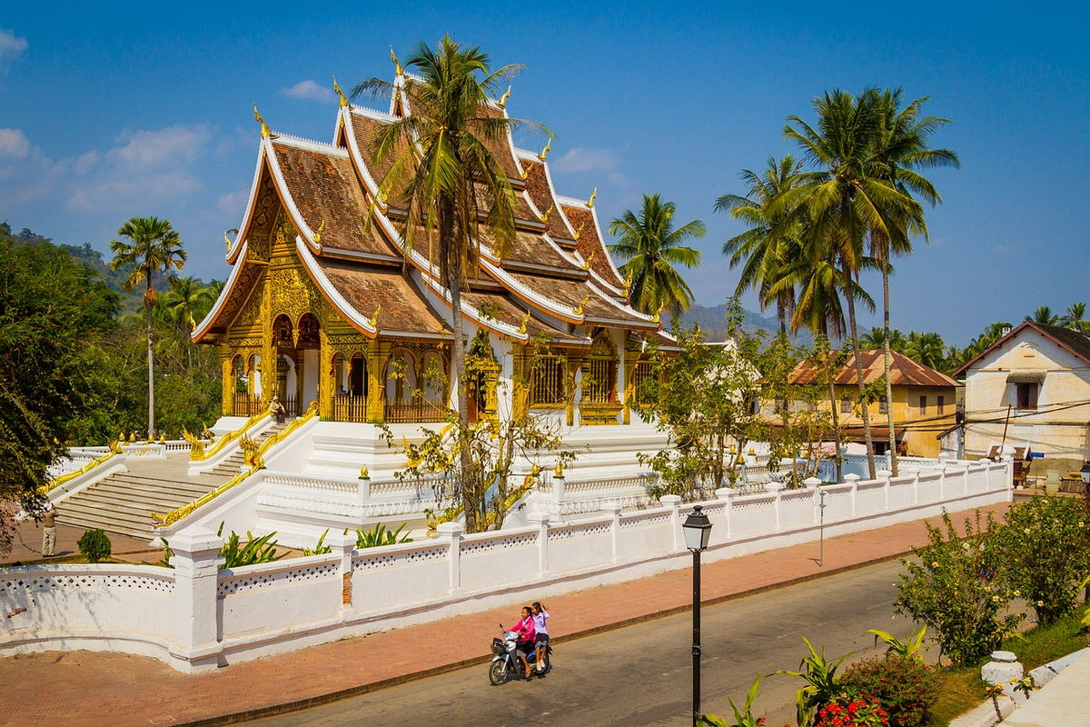 Un temple thaïlandais avec des toits en pente ornés de dorures, entouré d'arbres palmistes et d'une clôture blanche, avec deux personnes à moto en premier plan.