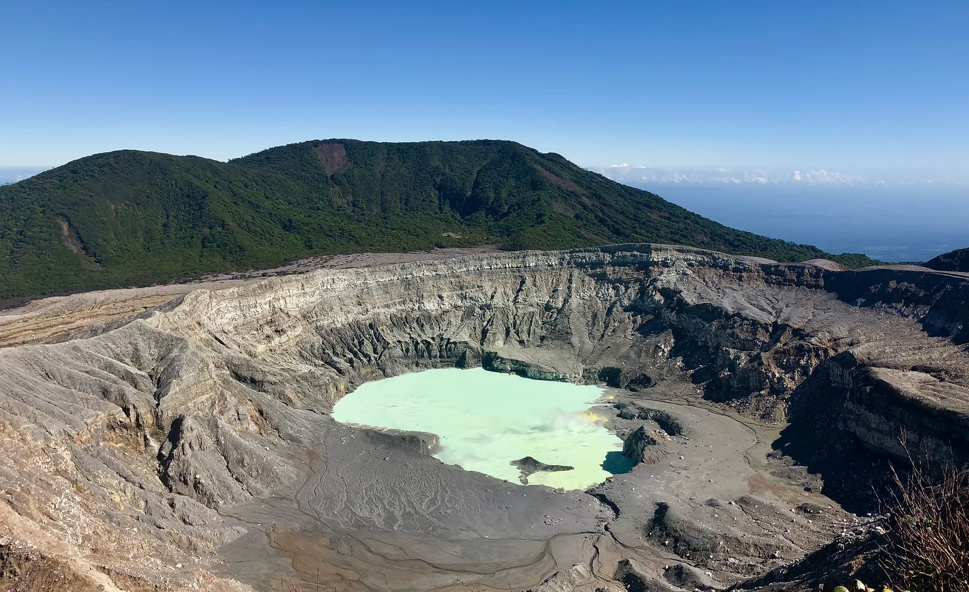 Vue d'un cratère de volcan avec un lac de couleur turquoise au centre, entouré de parois rocheuses et une forêt dense au sommet des montagnes en arrière-plan, sous un ciel bleu clair.