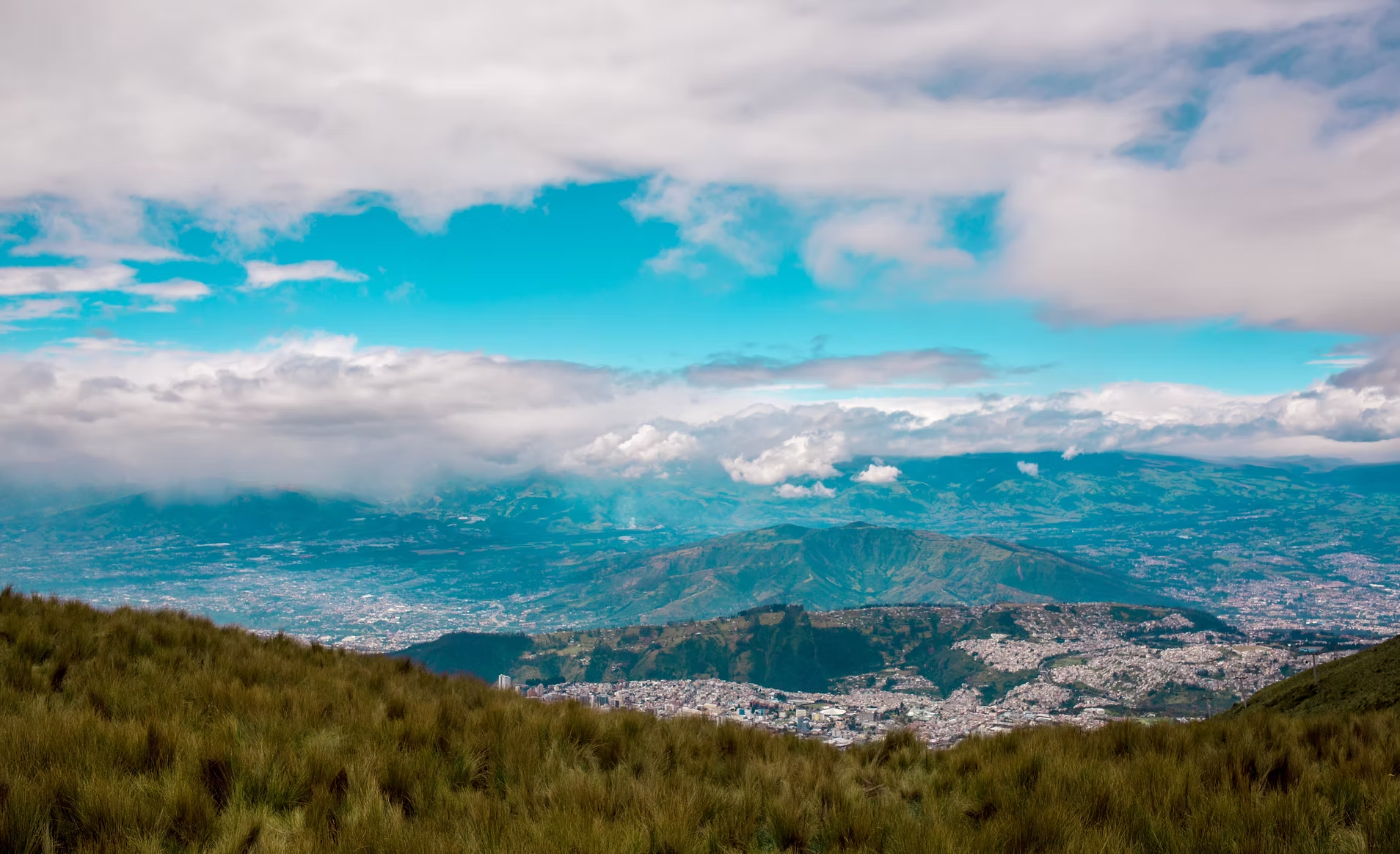 Vue panoramique sur une vallée entourée de montagnes sous un ciel partiellement nuageux, avec des zones de verdure au premier plan.