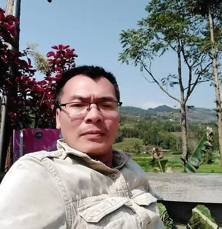Un homme porte des lunettes et une veste beige, assis à l'extérieur avec un paysage rural en arrière-plan, comprenant des arbres, des champs verts et des montagnes sous un ciel bleu clair.