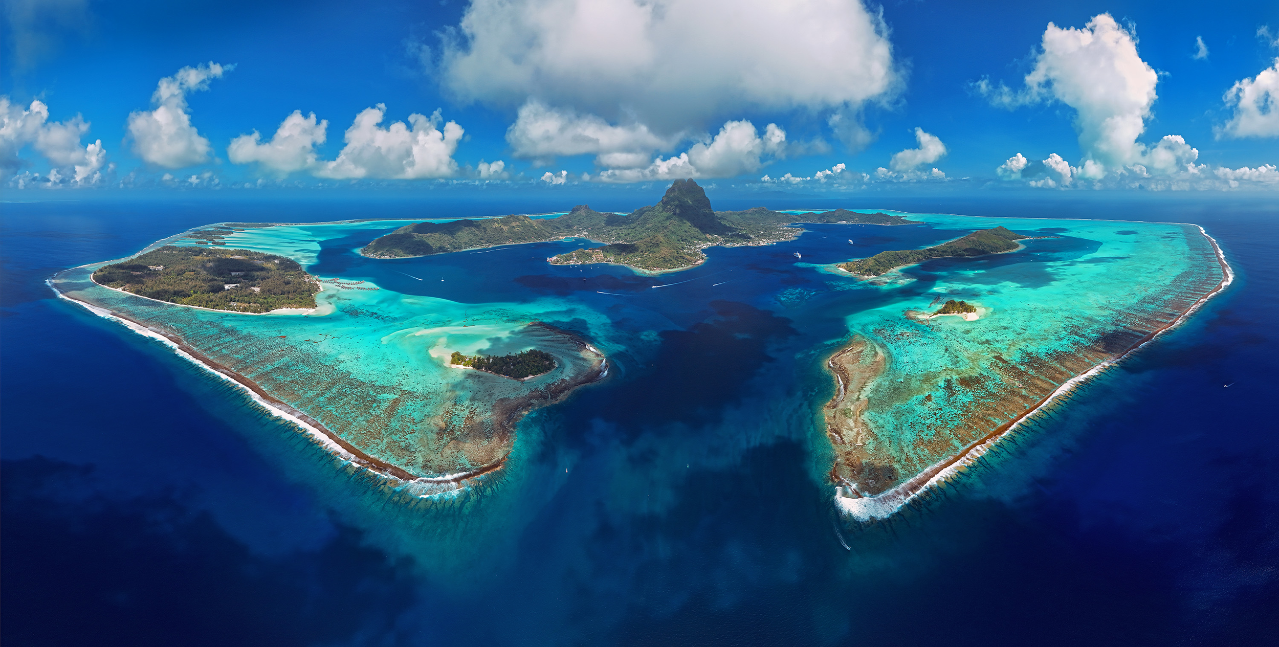 Vue aérienne d'une île avec des eaux turquoise, un lagon et des plages de sable blanc, entourée par l'océan bleu profond, avec quelques nuages dans le ciel.
