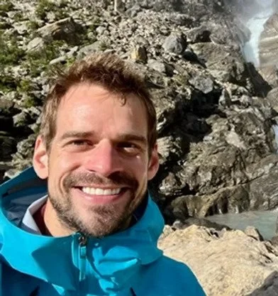 Un homme souriant prend un selfie devant une cascade rocheuse.