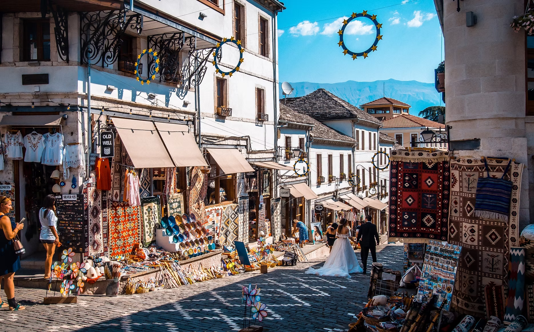 une rue animée avec des boutiques vendant des textiles, des souvenirs et des objets artisanaux, avec des personnes qui se promènent, un ciel bleu avec quelques nuages, des montagnes en arrière-plan, et des décorations circulaires suspendues dans la rue