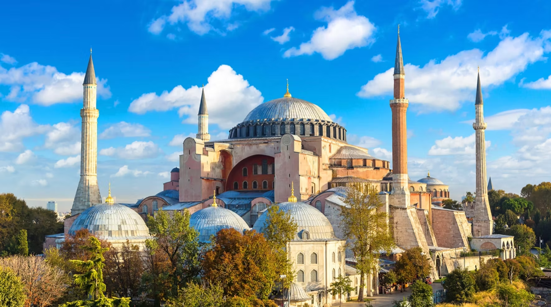 Vue de la mosquée Sainte-Sophie avec ses domes et ses minarets, entourée d'arbres colorés, le ciel bleu avec quelques nuages.