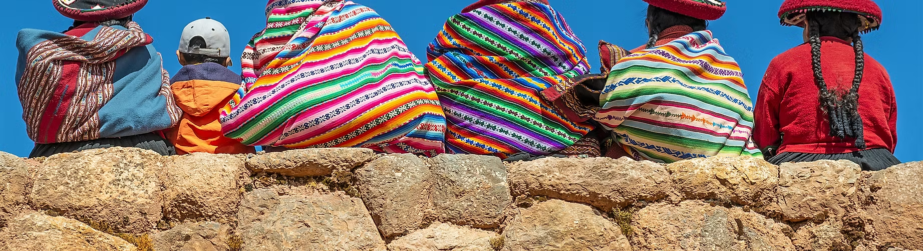 Groupe de personnes portant des vêtements traditionnels colorés, assises sur un mur en pierre, avec un ciel bleu en arrière-plan.