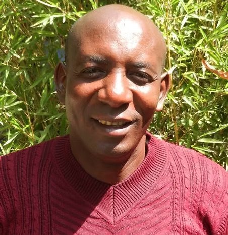 Un homme souriant portant un pull rouge devant une végétation verte dense.
