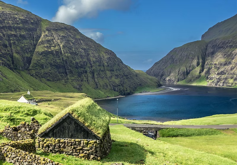 Paysage de montagnes verdoyantes entourant un lac bleu, avec quelques maisons traditionnelles en pierre et un ciel ensoleillé