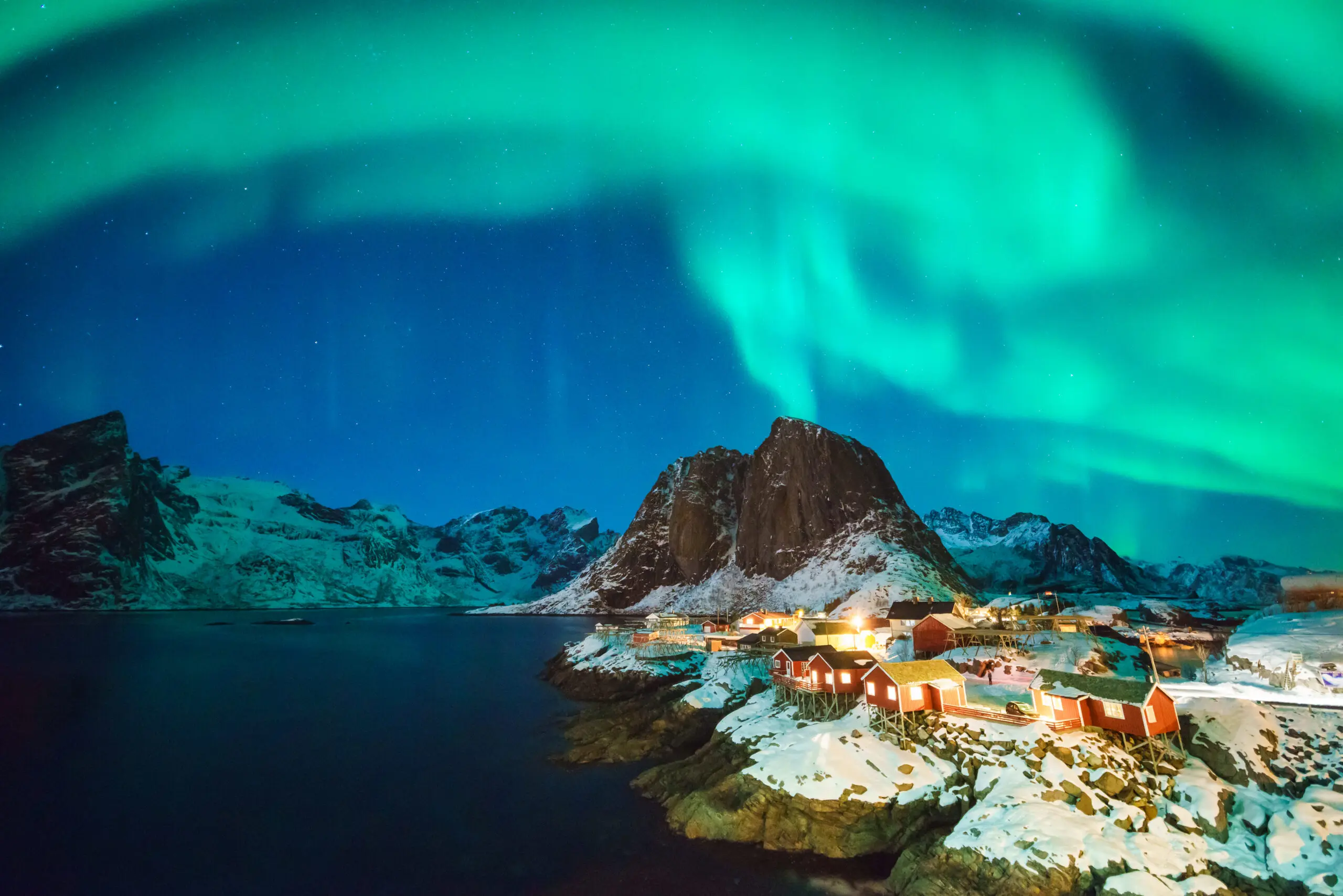 Aurore boréale dans un ciel étoilé avec des montagnes enneigées et un village au bord d'un fjord.