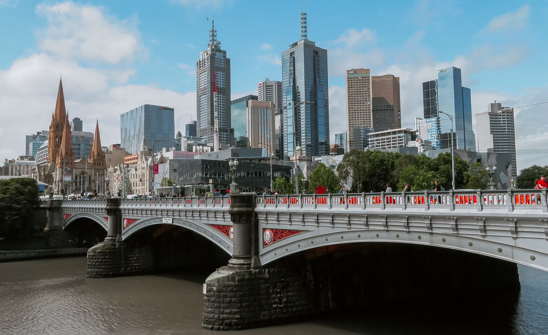 Vue d'une ville avec des bâtiments modernes et une église en architecture gothique, un pont en bois avec des piétons, et des arbres le long de la rivière sous un ciel partiellement nuageux.