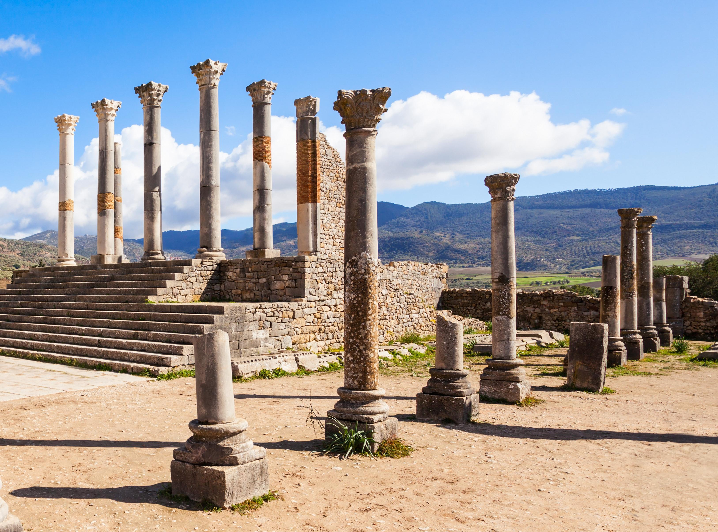 Restes de colonnes antiques sur des escaliers en pierre dans un site archéologique, avec un paysage montagneux en arrière-plan, sous un ciel bleu avec des nuages.