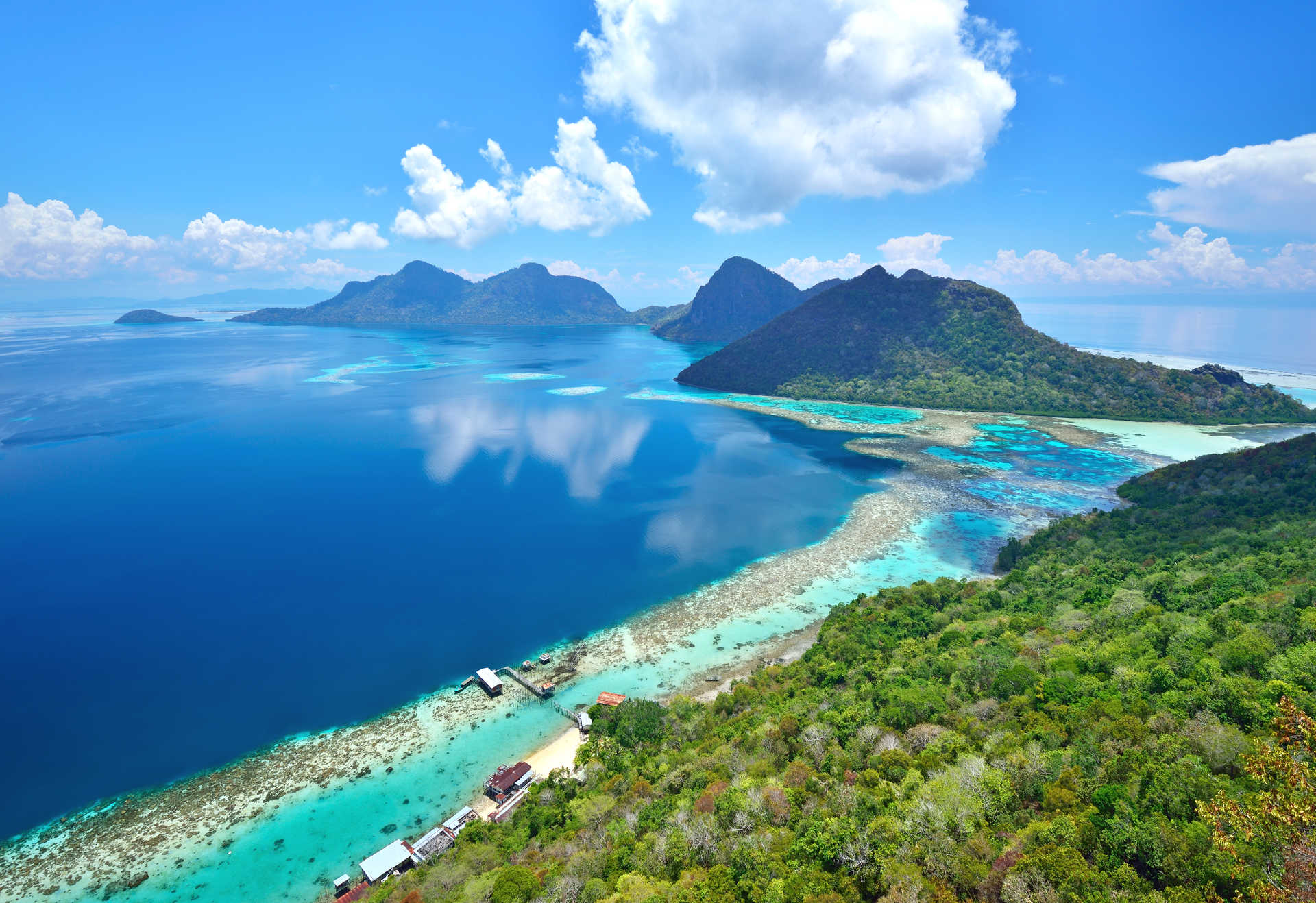 Vue aérienne d'îles tropicales avec montagnes, eaux turquoise, et forêt dense. Cabanes et ponton au bord de l'eau.