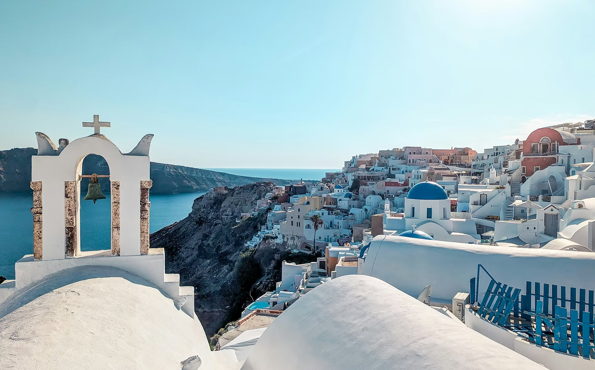 Vue de Santorin, avec une église blanche à clocher et une croix, en arrière-plan des maisons blanches et des toits bleus sur une falaise surplombant la mer Égée, sous un ciel clair.