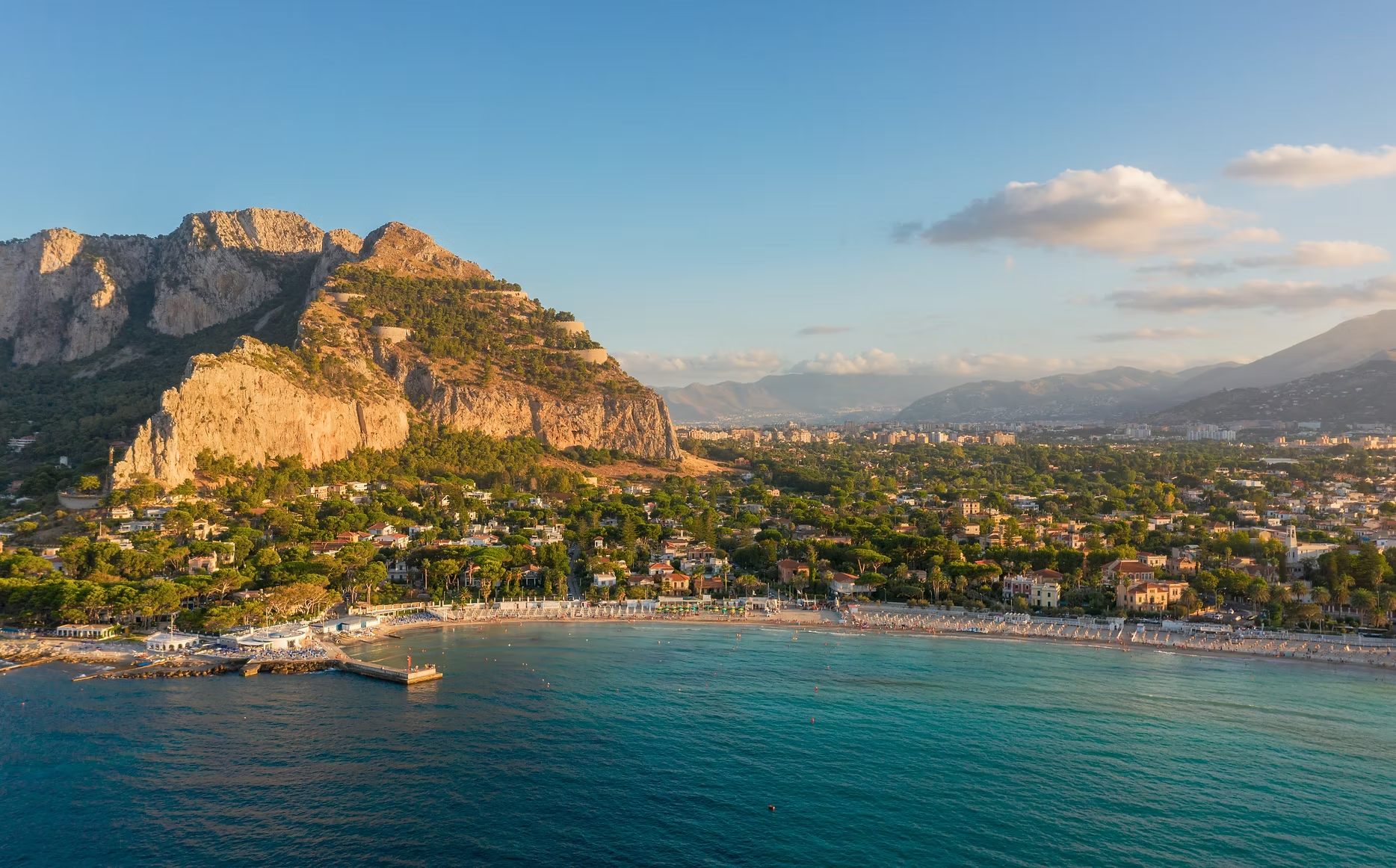 Vue panoramique d'une ville côtière avec une plage, des bâtiments résidentiels, entourée de montagnes et de collines avec un ciel en partie nuageux.