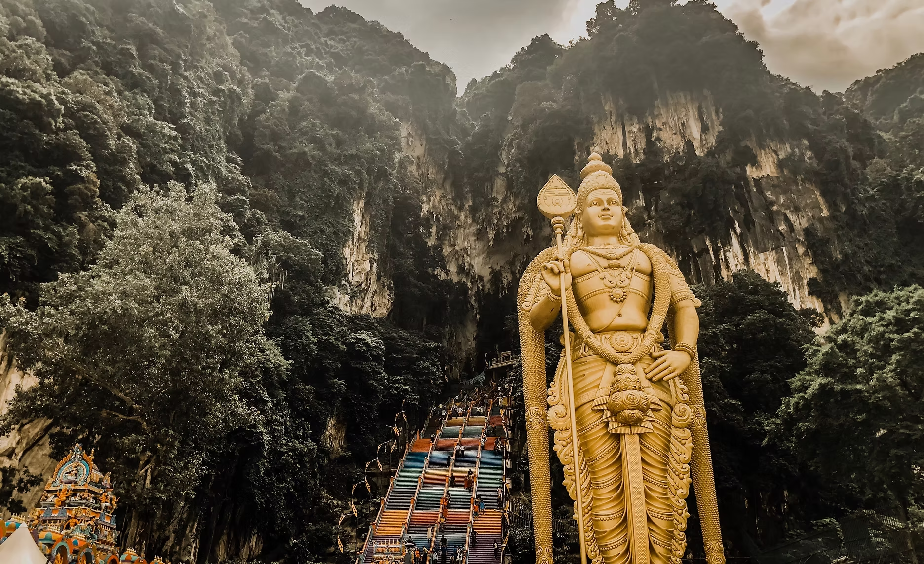 Giant statue de Hanuman au pied de la grotte de Batu, avec escalier coloré menant à l'entrée de la grotte, entourée de végétation dense et de formations rocheuses.