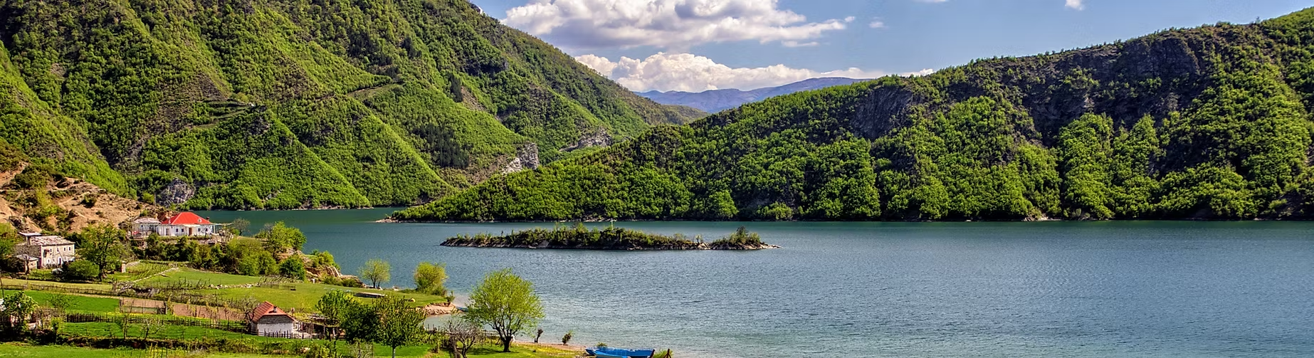 Paysage de lac entouré de montagnes verdoyantes avec quelques maisons en dessous et un petit bateau sur l'eau.
