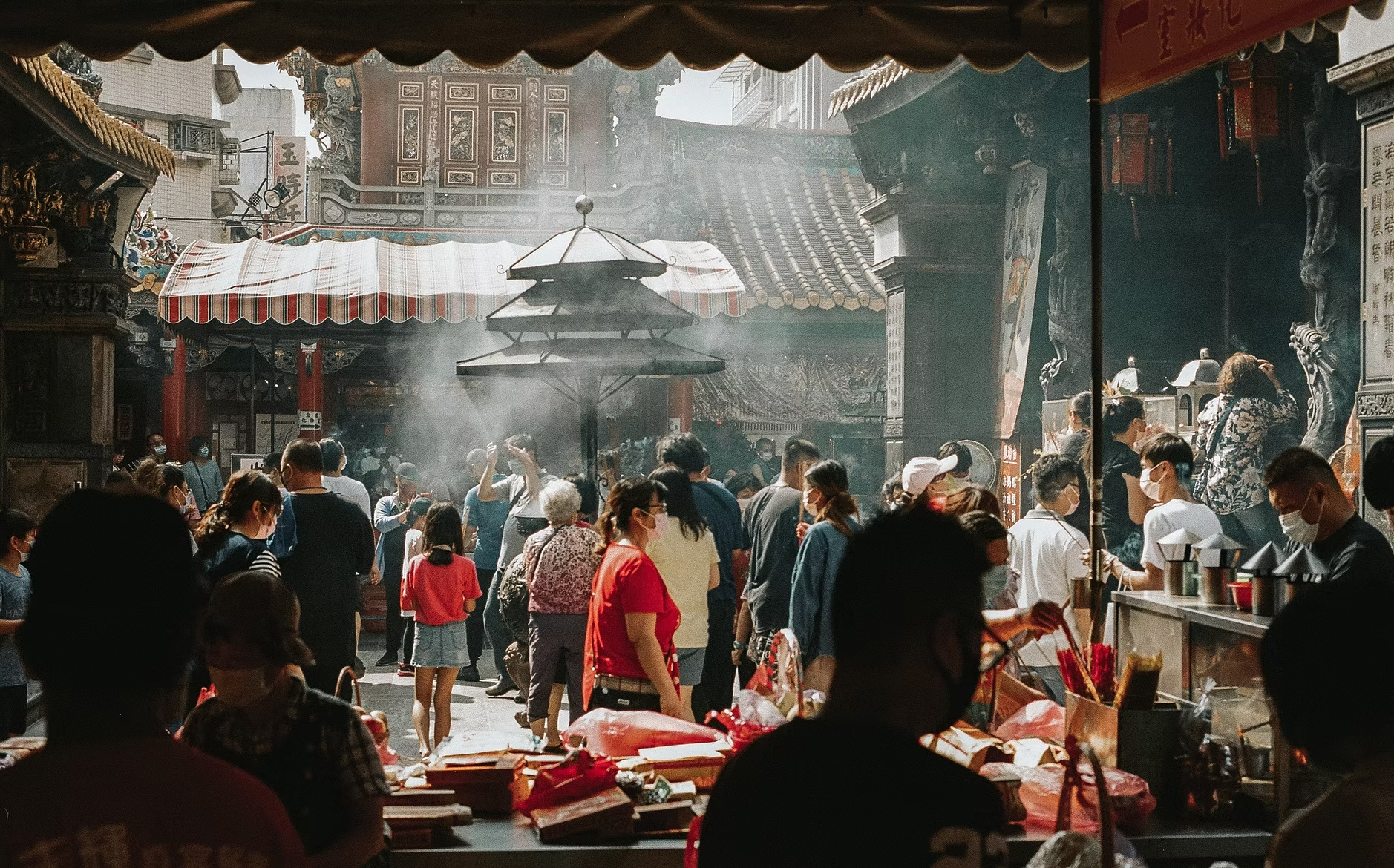 Vue d'un marché ou d'un temple asiatique bondé, avec des personnes portant des masques, sous une structure couverte traditionnelle, avec des temples et des enseignes en arrière-plan.