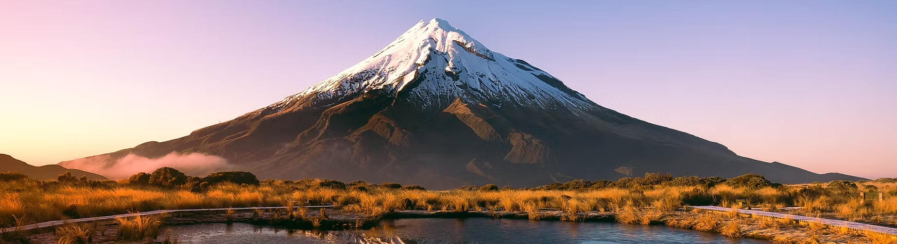 Un paysage naturel avec un volcan enneigé au sommet, entouré d'une plaine herbeuse avec un étang ou un lac au premier plan, sous un ciel clair au coucher du soleil.