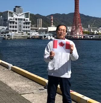 Homme tenant un drapeau du Canada devant un corps d'eau et une tour Rouge avec des montagnes en arrière-plan.