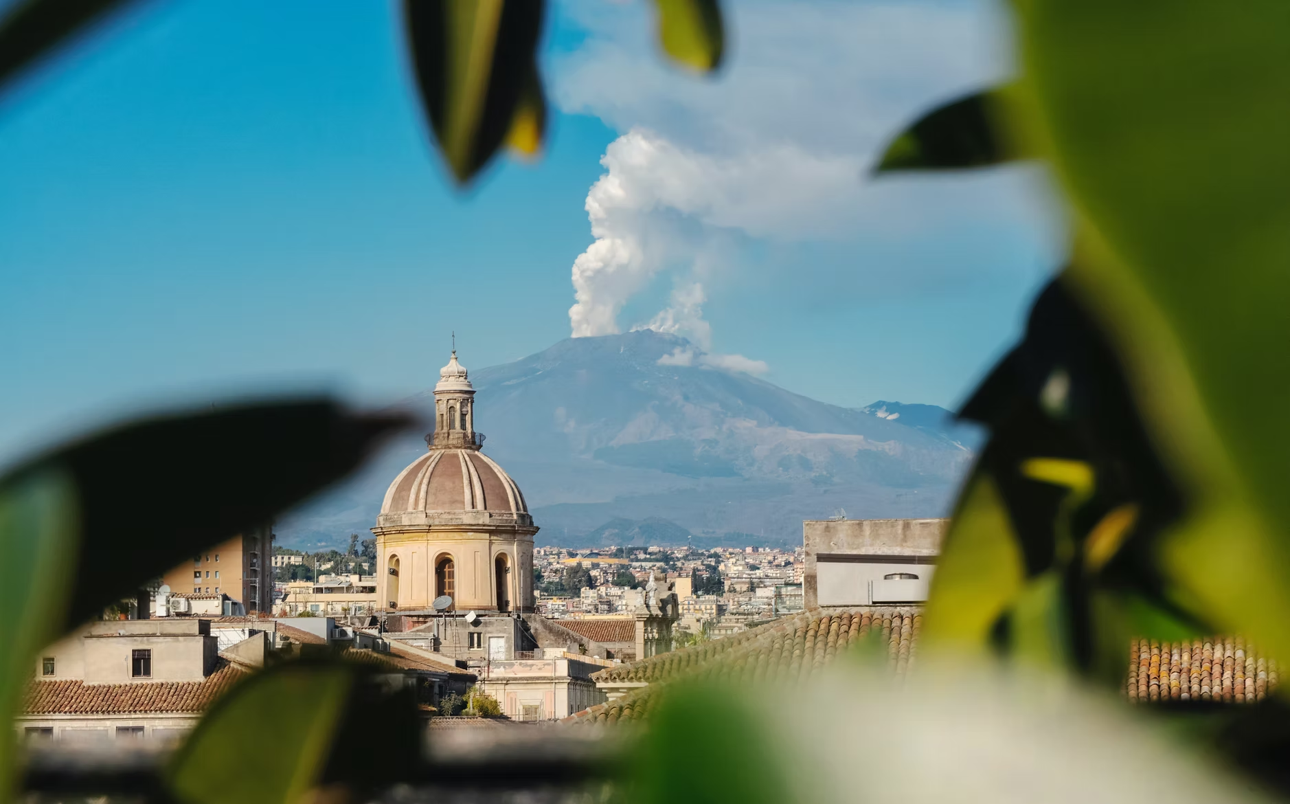 Vue d'une ville avec une église en premier plan, un volcan en arrière-plan, encadrée par des feuilles vertes.