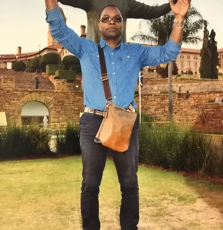 Un homme portant une chemise bleue et un sac à bandoulière posé en travers du torse, se tenant debout dans un parc avec des bâtiments historiques en arrière-plan.