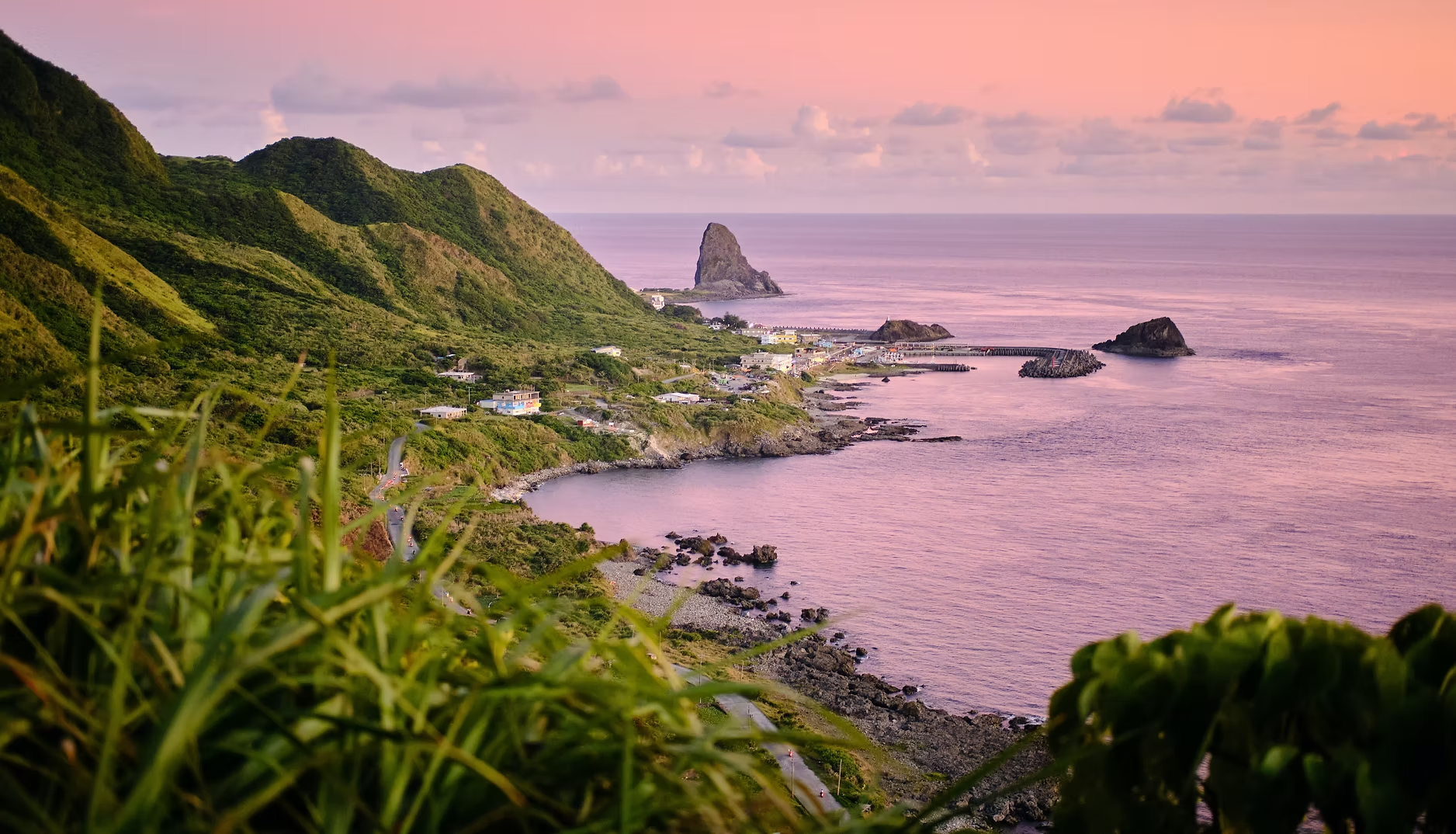 Paysage côtier avec montagnes verdoyantes, petits bâtiments, et mer sous un ciel rose au coucher du soleil.
