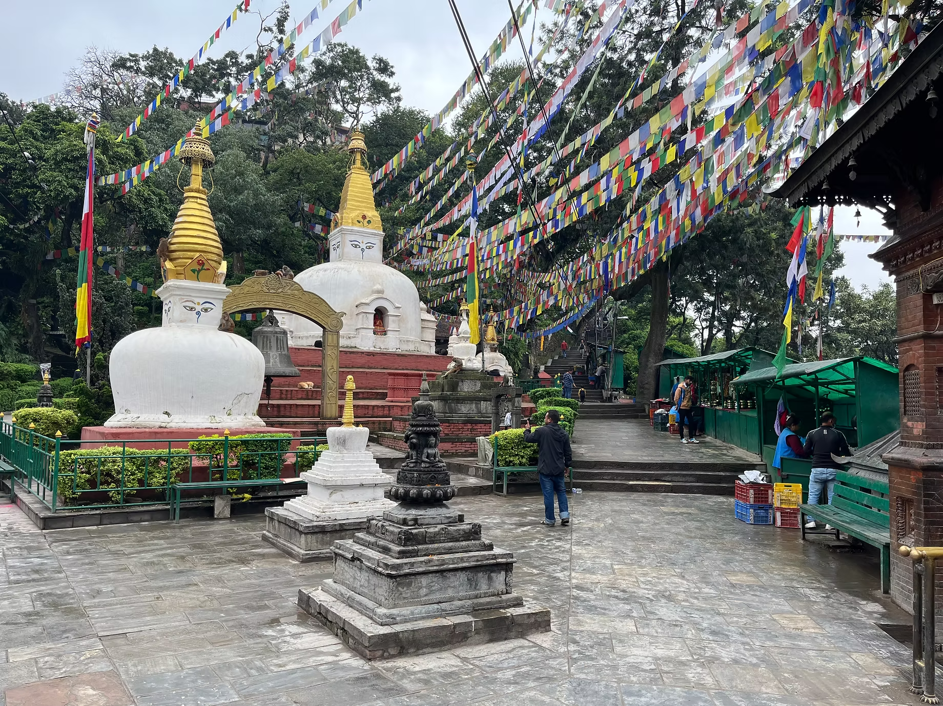 Temple bouddhiste avec des drapeaux colorés suspendus au-dessus, statues de Bouddha et personnes se promenant dans la cour pavée, arbres en arrière-plan.