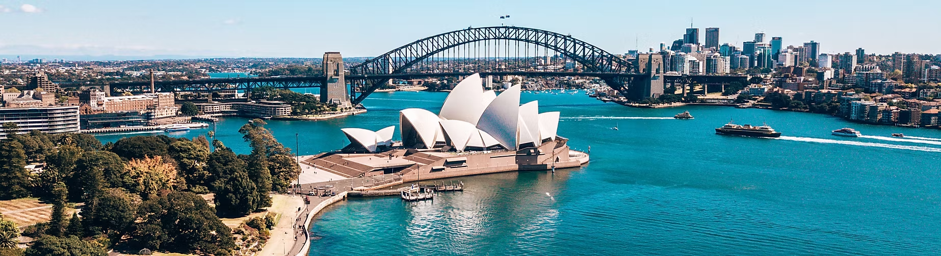 Vue panoramique de la baie de Sydney avec l'Opéra de Sydney au centre, le Harbour Bridge en arrière-plan, et la ville de Sydney avec ses gratte-ciel au loin.