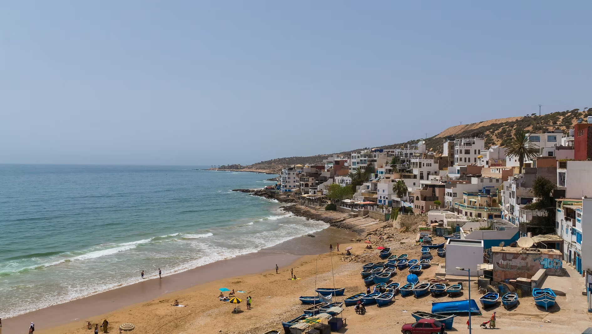 Plage avec des bateaux de pêche alignés, sable doré, habitants se promenant, maisons colorées en haut d'une colline, ciel clair et mer calme.