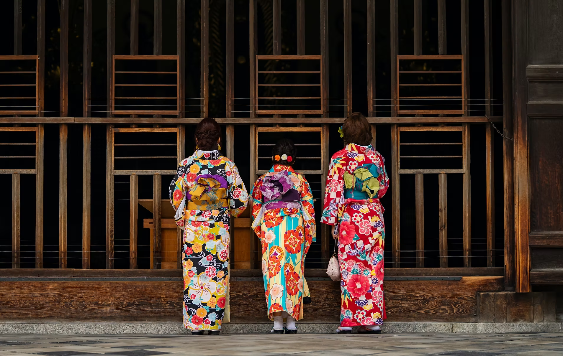 Trois femmes portant des kimonos traditionnels japonais avec des motifs floraux, allant de gauche à droite, se tenant devant une structure en bois.