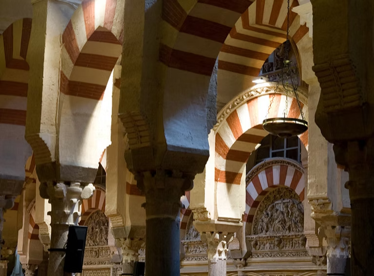 Intérieur d'une architecture mauresque avec des arches en pierre, rayures rouges et blanches, et des ornements sculptés, éclairé par un lustre suspendu.