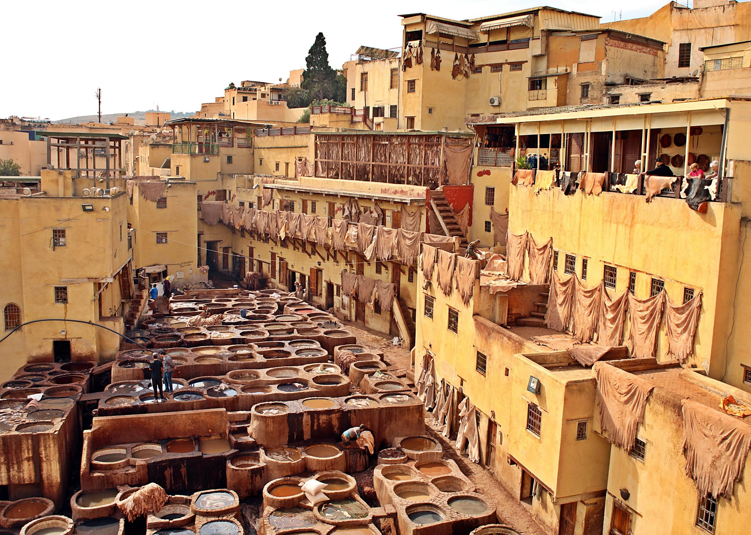 Une vue d'une ville berbère avec des bâtiments en terre cuite, des logements en hauteur et des terrasses, où des habitants sèchent des vêtements aux balcons, et des tanneries avec de grands récipients en pierre contenant des teintures ou des matières animales.