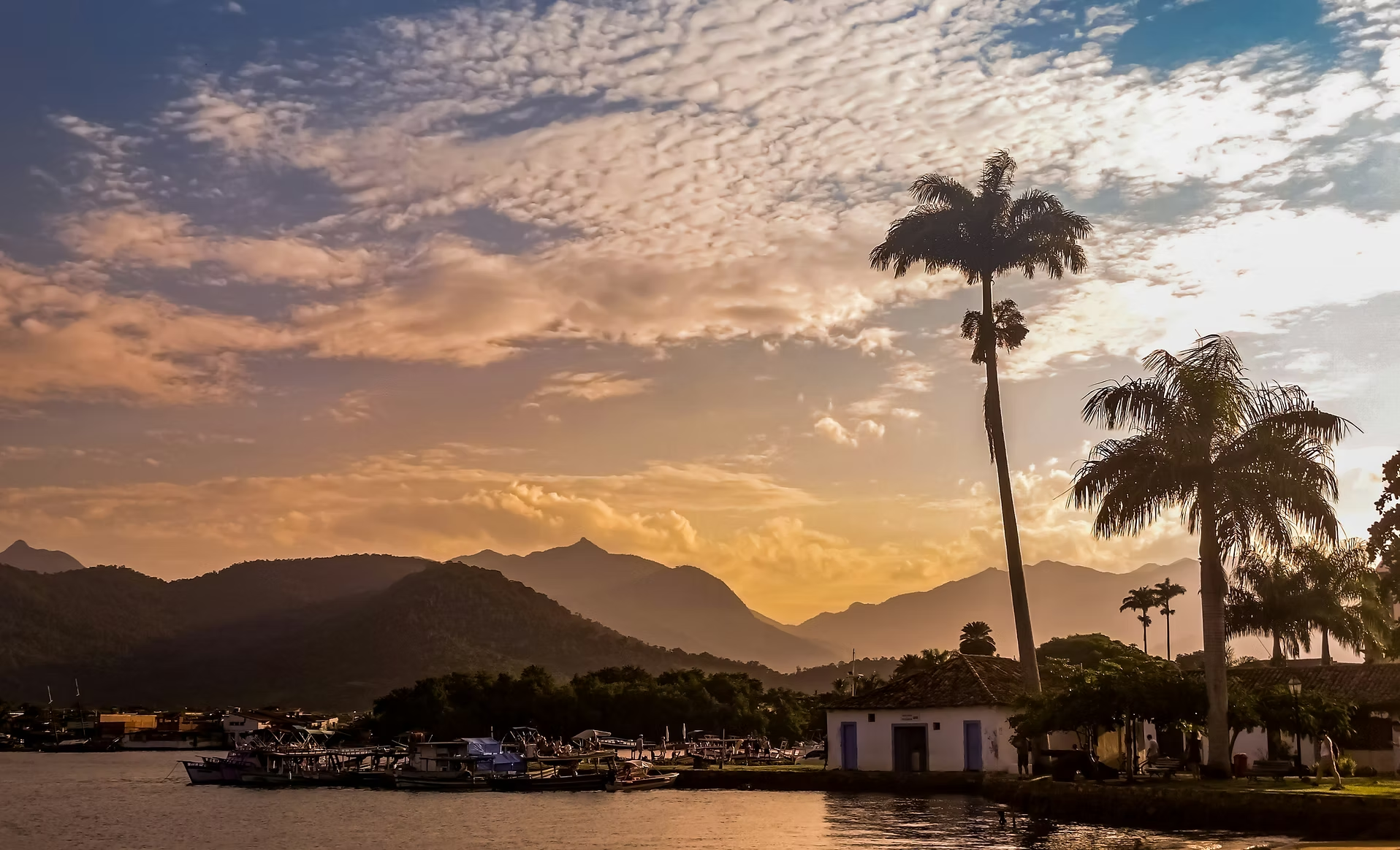 Coucher de soleil sur un paysage naturel avec des palmiers, des montagnes en arrière-plan et un lac avec des bateaux.