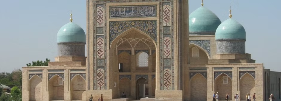 Vue de l'entrée principale d'une mosquée avec deux grandes coupoles turquoise et plusieurs petites, décorée de motifs géométriques et calligraphies, avec des personnes devant.