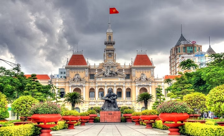 Une photo de l'hôtel de ville de Ho Chi Minh-Ville en Thaïlande, avec un monument au centre, des arbres verts et un ciel nuageux en arrière-plan.