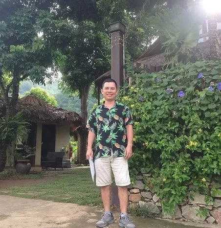 Un homme souriant en chemise à motif tropical, shorts beige, chaussures de sport, dans un jardin avec des arbres, des fleurs violettes et une maison en arrière-plan.