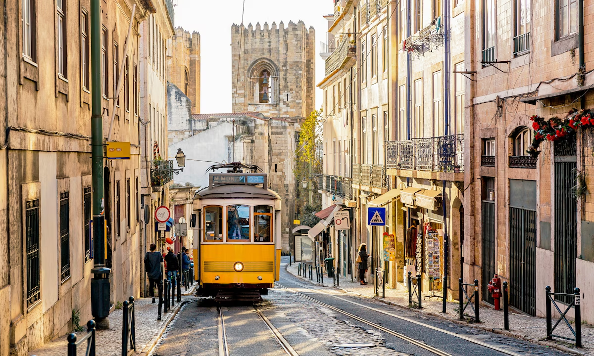 Un tram jaune circule dans une rue pavée bordée de bâtiments anciens, avec une église en pierre en arrière-plan, à Lisbonne, Portugal.