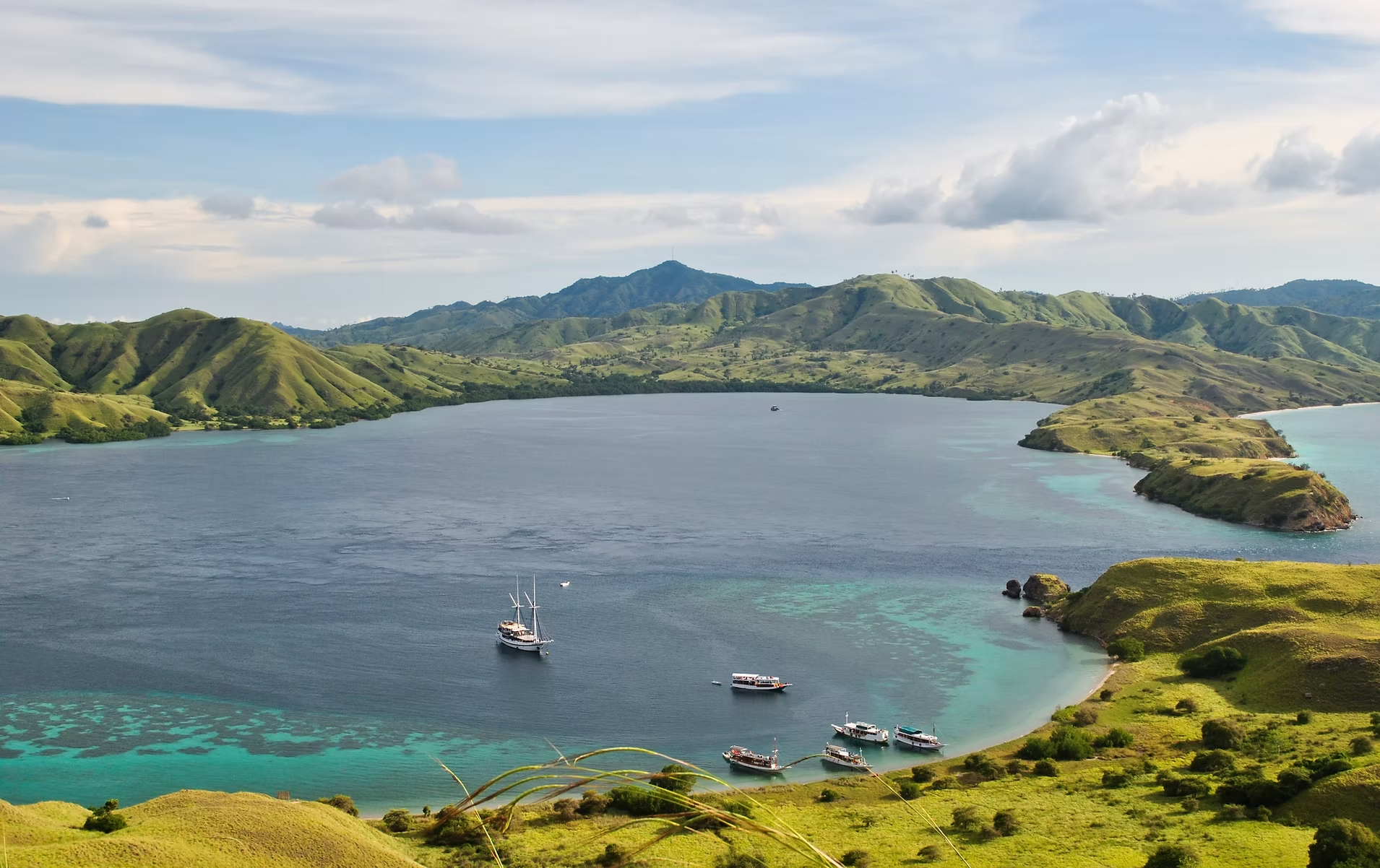 Paysage avec une grande étendue d'eau entourée de collines vertes et de montagnes au loin, avec quelques bateaux à quai.