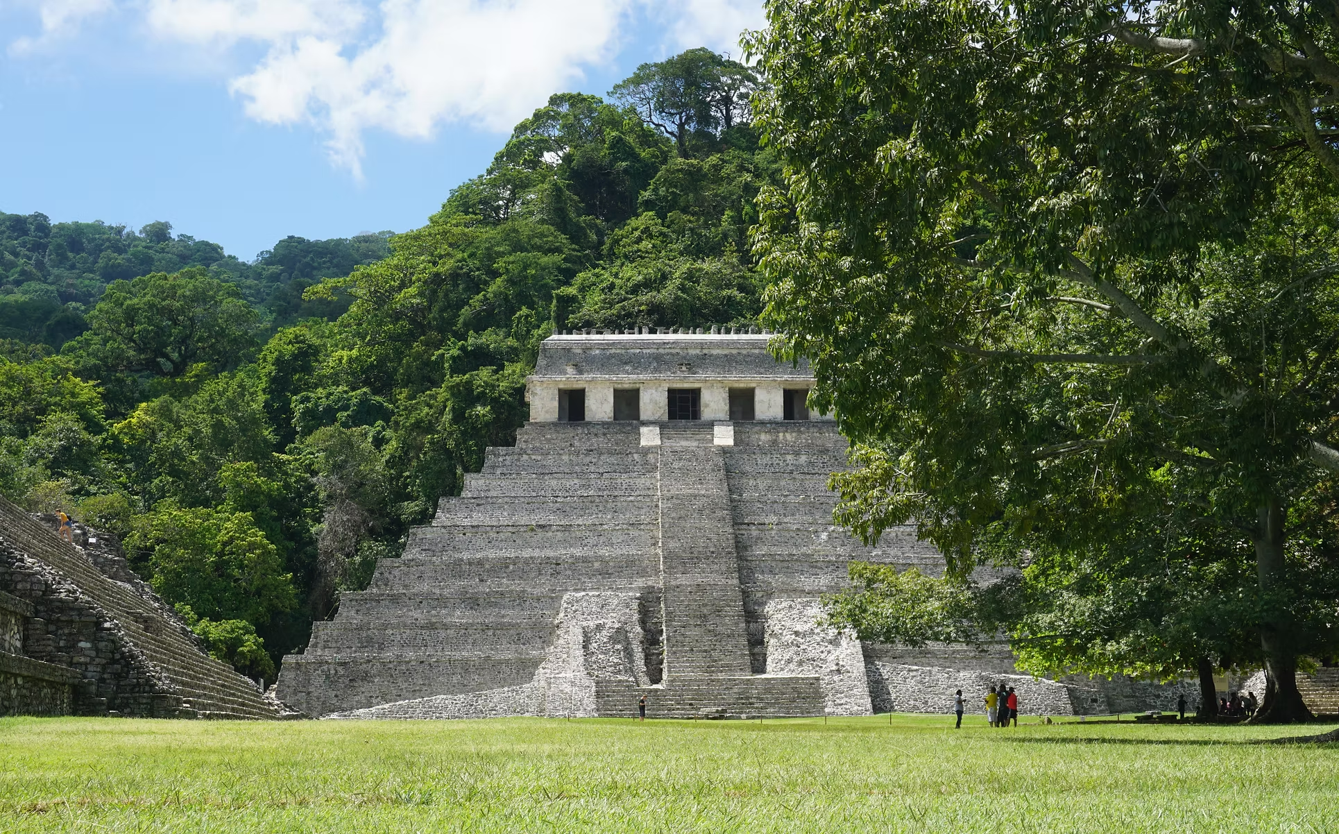 Vue d'une ancienne pyramide maya en pierre dans une forêt dense, avec des escaliers, sous un ciel partiellement nuageux, entourée d'arbres verts