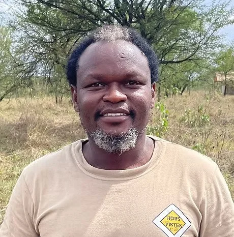 Homme afro-descendant portant un t-shirt beige avec un logo, en extérieur avec des arbres en arrière-plan.