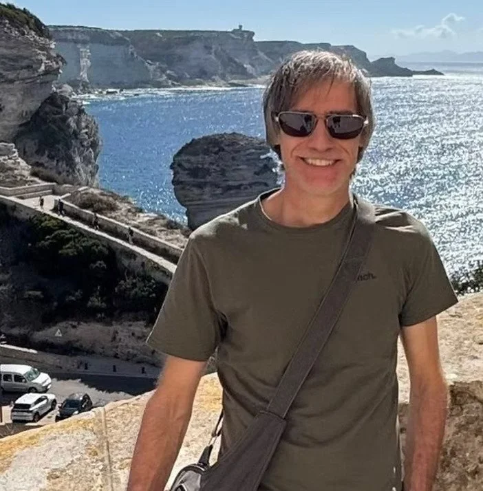 Un homme souriant portant des lunettes de soleil, une chemise verte, et une sacoche, se tient près de la côte avec la mer, des falaises et un ciel en arrière-plan.