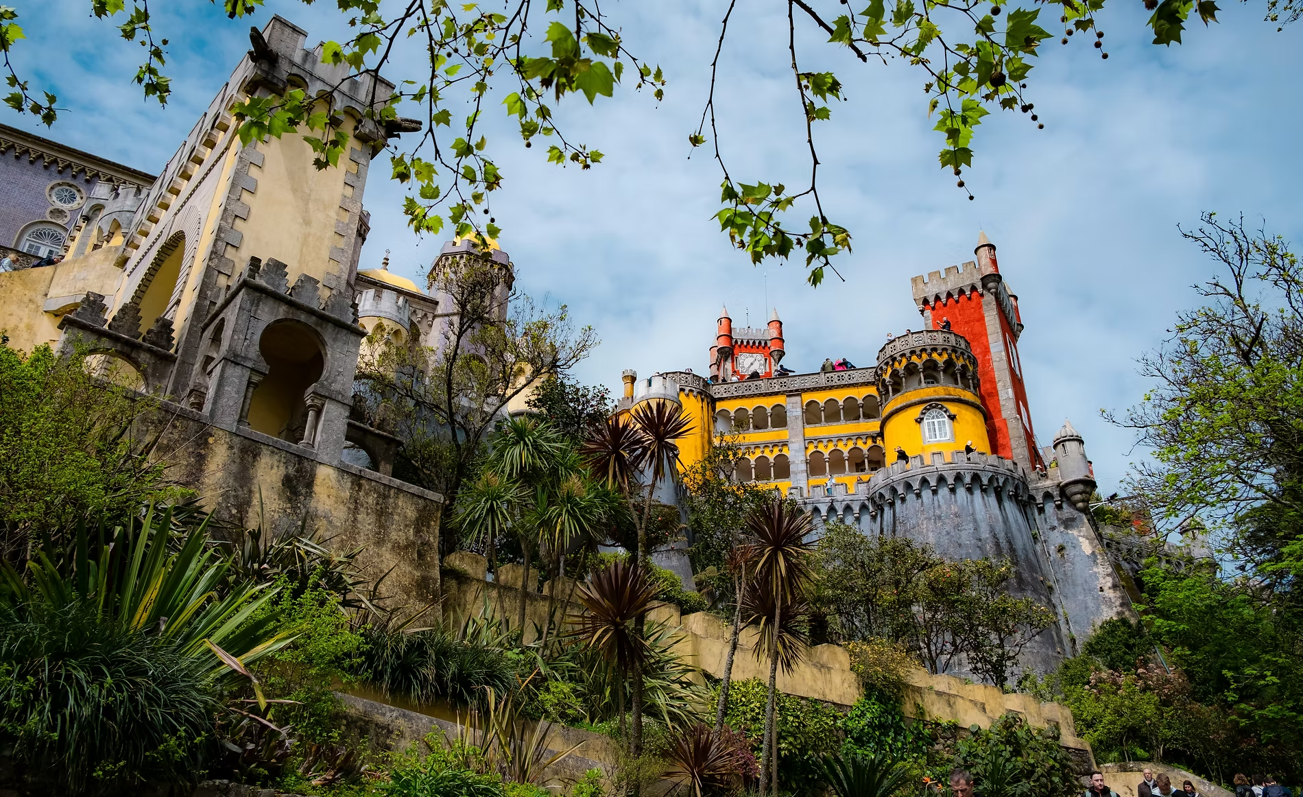 Château coloré en style médiéval, avec des tours et des murailles, entouré de végétation luxuriante et d'arbres, sous un ciel partiellement nuageux.
