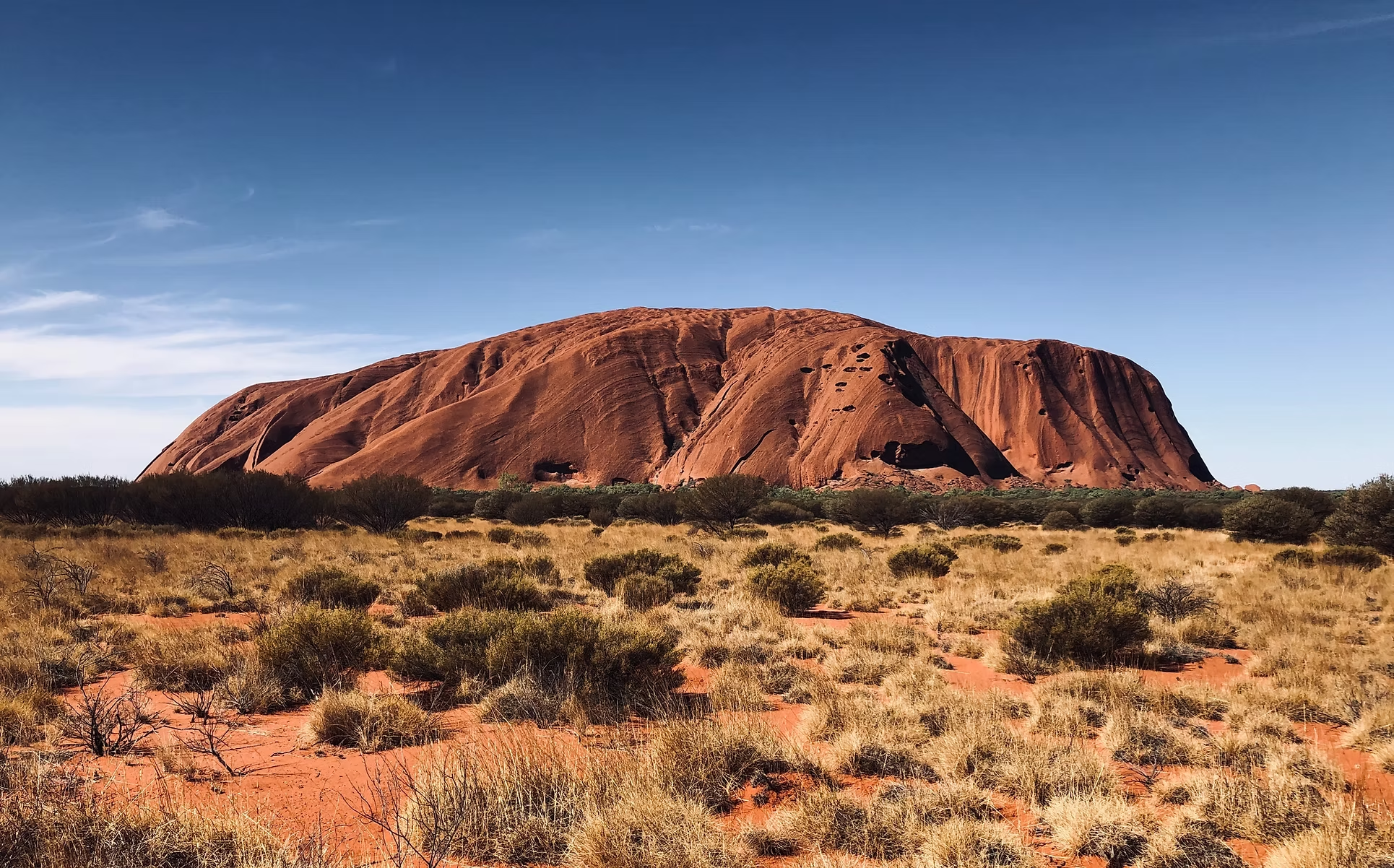 Une grande formation rocheuse orange dans un paysage de savane avec des petits arbustes et un ciel bleu clair.