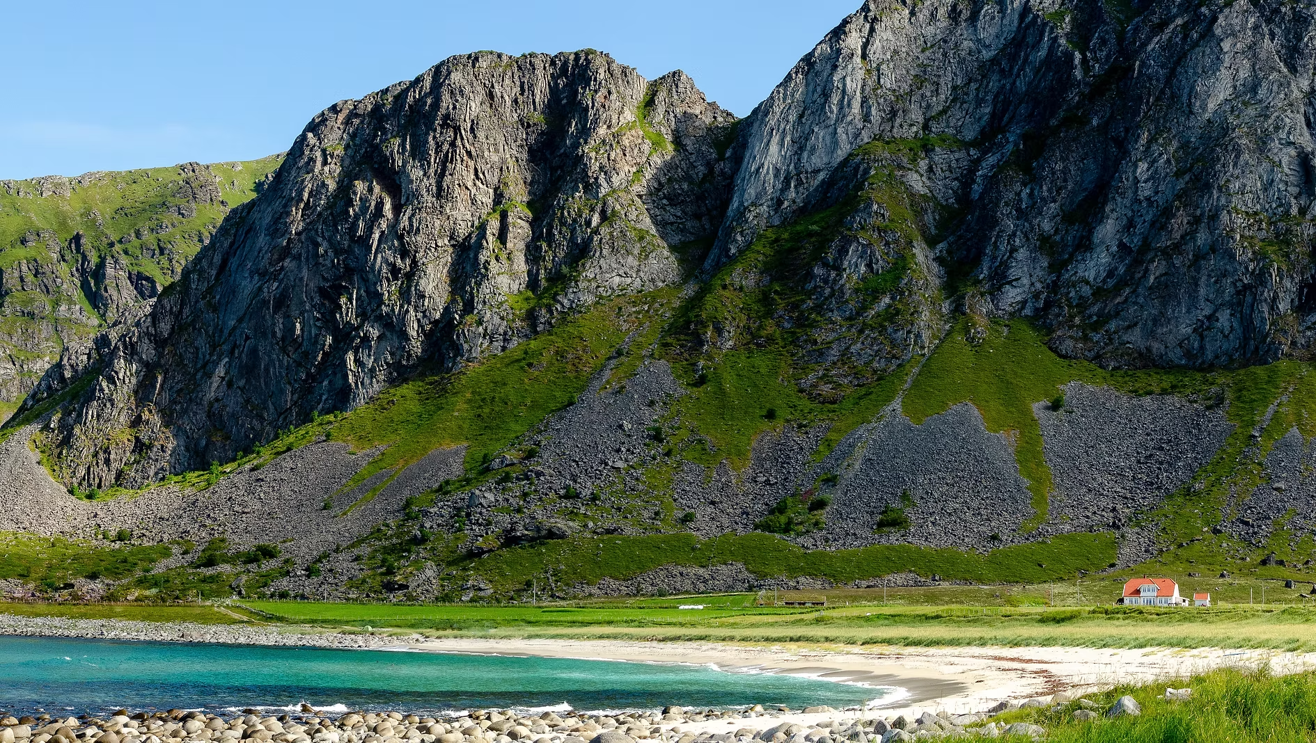 Vue d'une maison blanche avec un toit rouge au pied de montagnes rocheuses avec de la végétation verte et un lac en premier plan.
