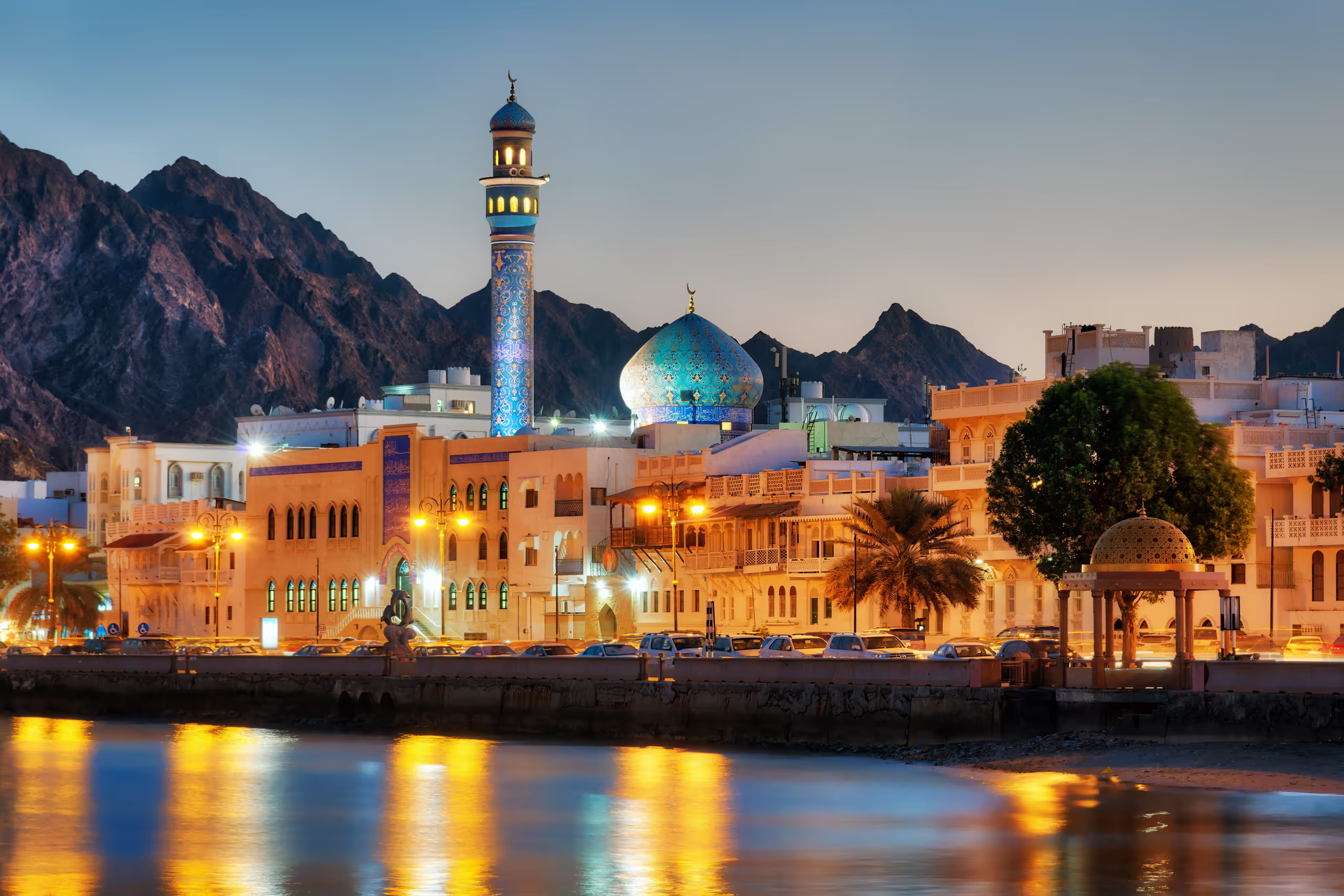 Mosquée avec dôme bleu et minaret illuminé, bâtiments traditionnels, montagnes en arrière-plan, reflection dans l'eau au premier plan, fin de journée ou début de soirée.