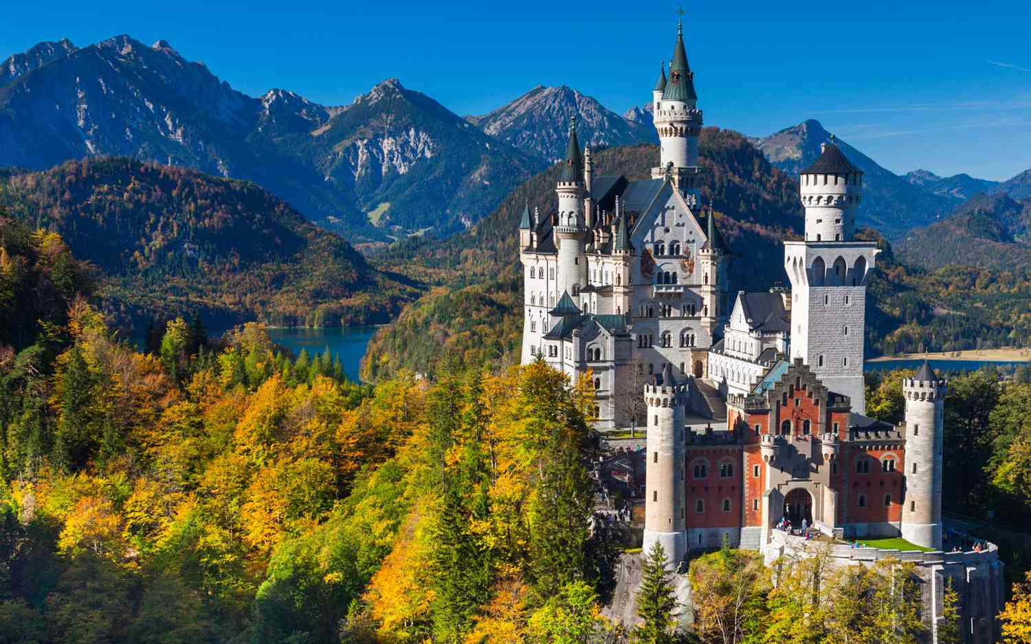 Château de Neuschwanstein entouré de forêts colorées et de montagnes en arrière-plan, sous un ciel bleu.