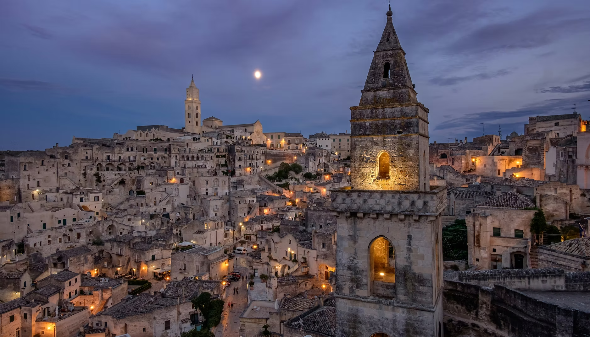 Vue panoramique de la ville de Matera en Italie, avec des bâtiments en pierres blanches, une église avec un clocher illuminé, sous un ciel nocturne avec la lune visible.
