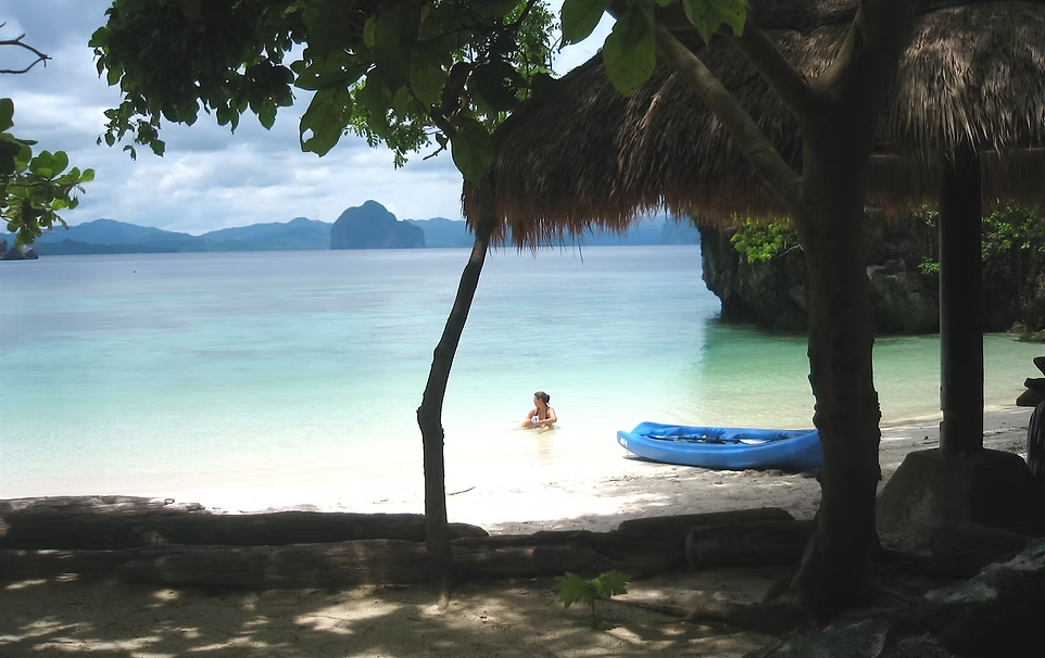 Plage tropicale avec eau turquoise, personne nageant, kayak bleu, huttes en toit de paille, arbres verts, montagnes en arrière-plan.