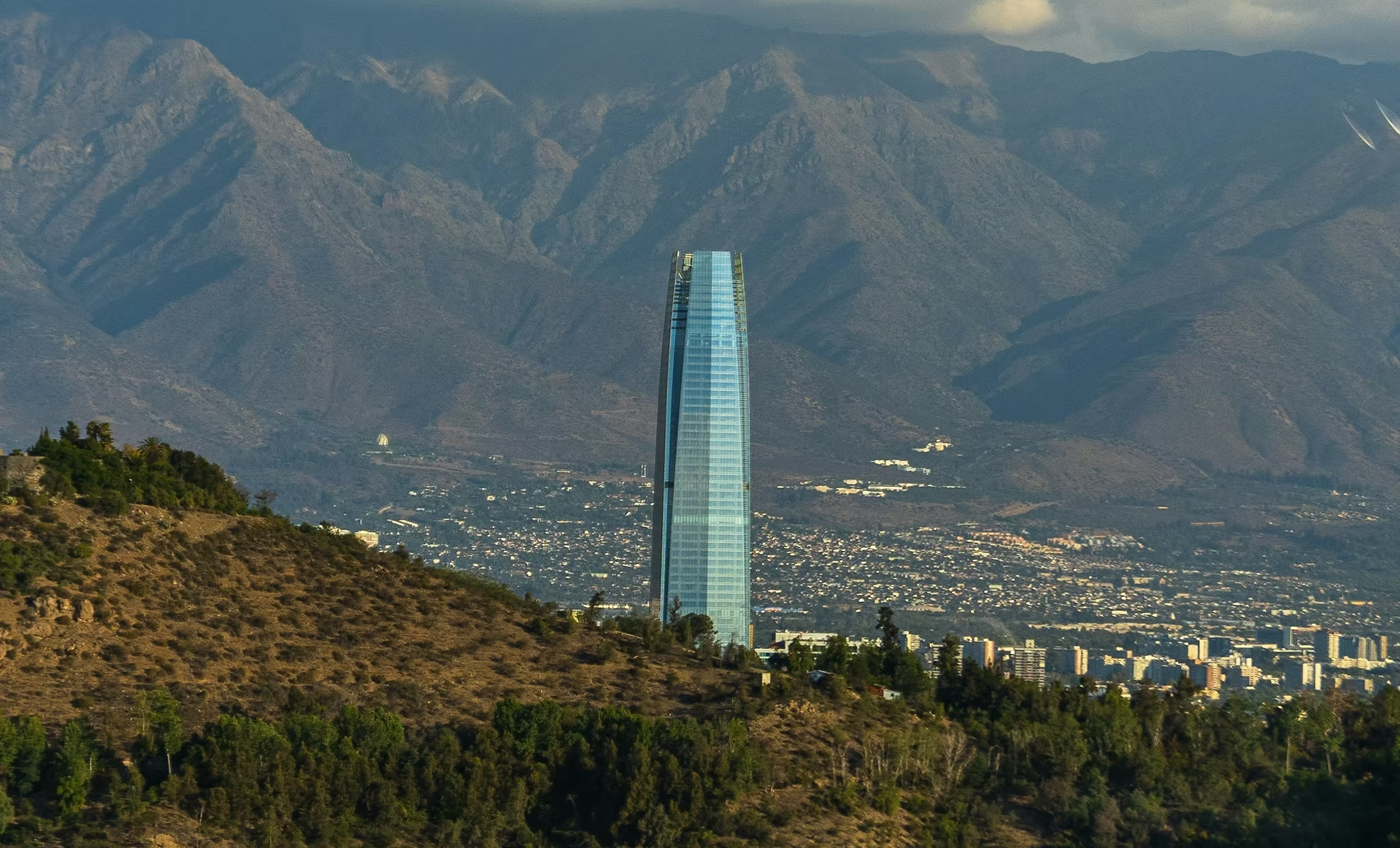 Gratte-ciel moderne en verre dans une ville au pied de montagnes rocheuses, avec une végétation aride au premier plan.
