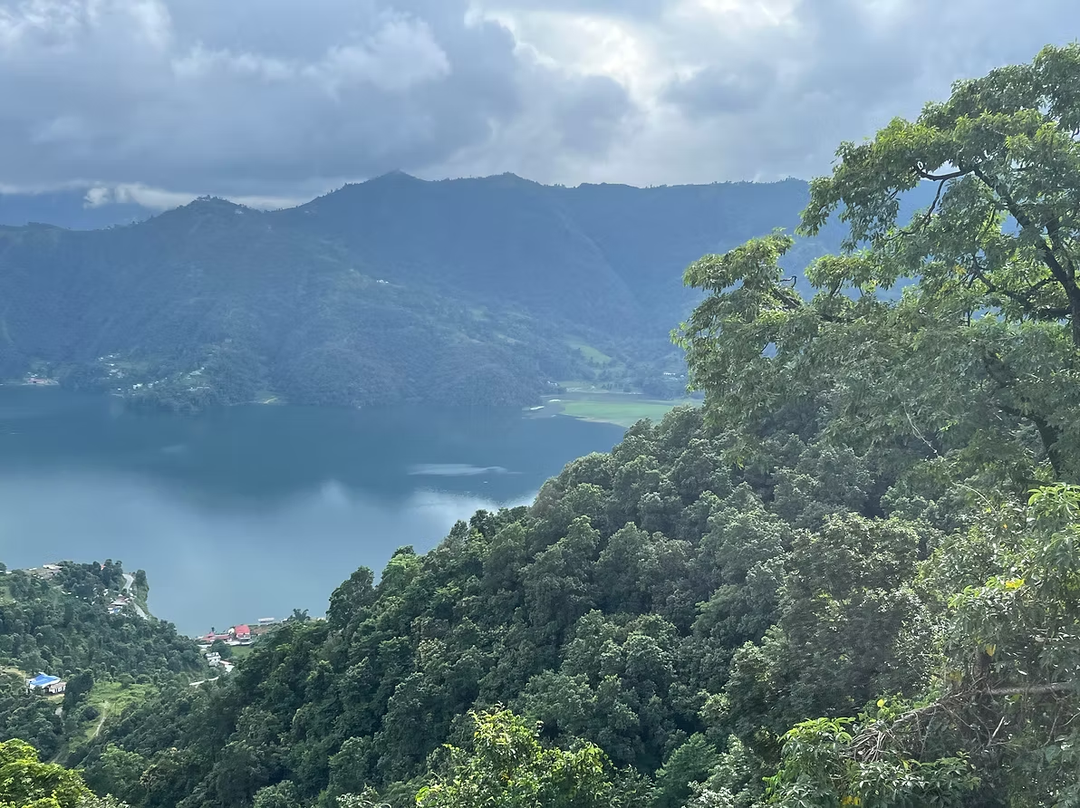 Vue panoramique d'un lac entouré de montagnes verdoyantes et de forêts denses, avec un ciel nuageux.