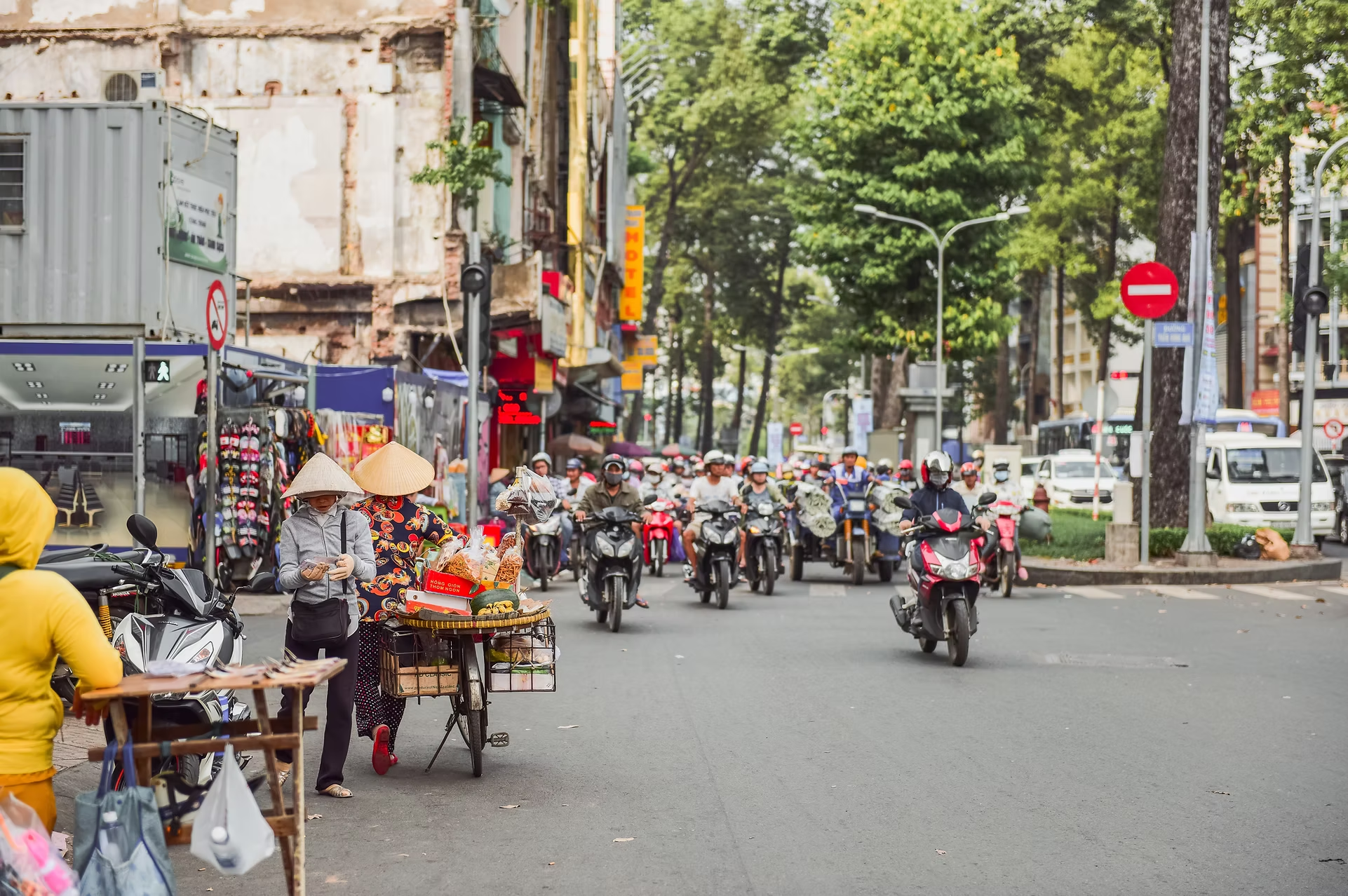 Rue animée avec des moto-taxis, une vendeuse avec un vélo marchand des produits, des personnes portant des chapeaux coniques, des magasins et des panneaux publicitaires. Plusieurs arbres et lampadaires bordent la rue.